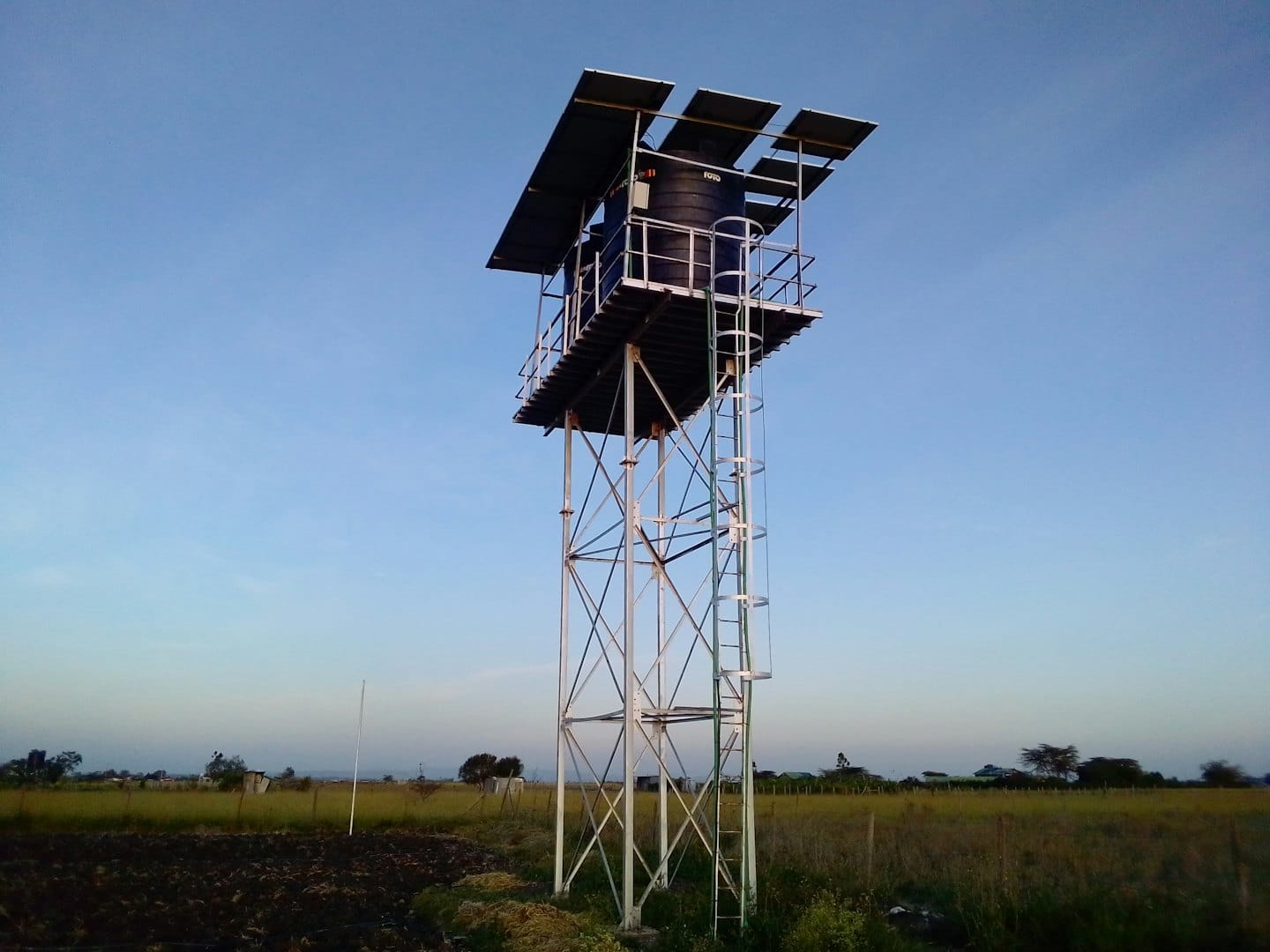 Metal water tower with solar panels stands in a field.